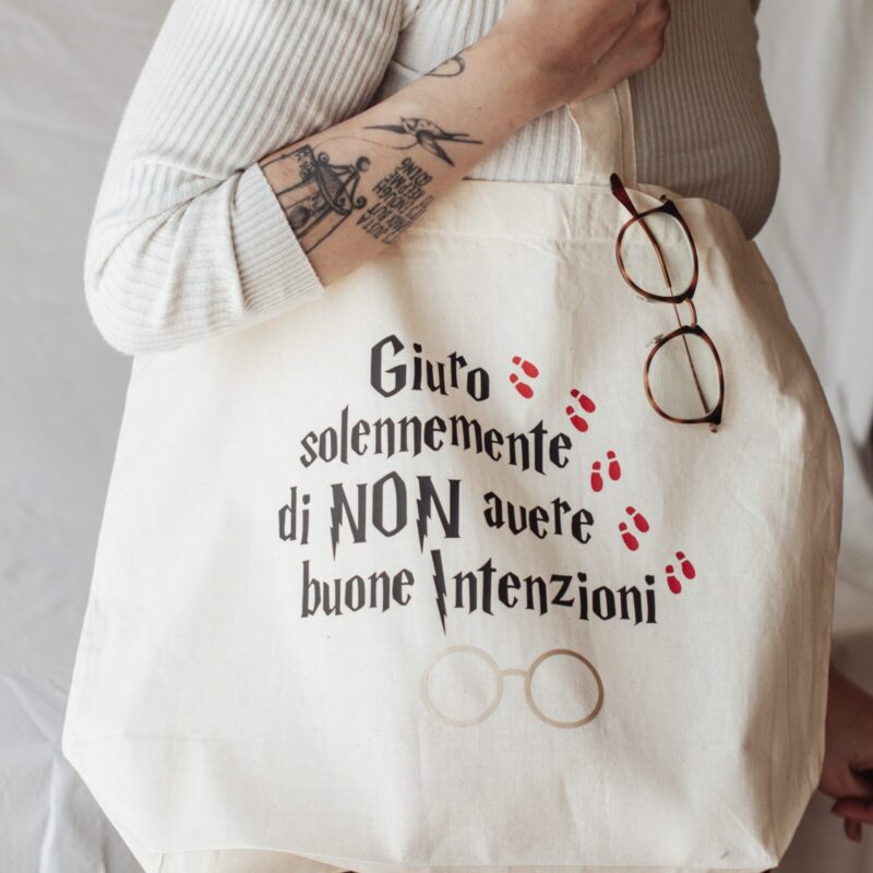 Ragazza che porta la Tote bag color beige con la scritta "Giuro solennemente di non avere buone intenzioni" con il font di Harry Potter in nero, cinque impronte rosse di scarpa in sequenza e il disegno degli occhiali di Harry Potter color dorato.